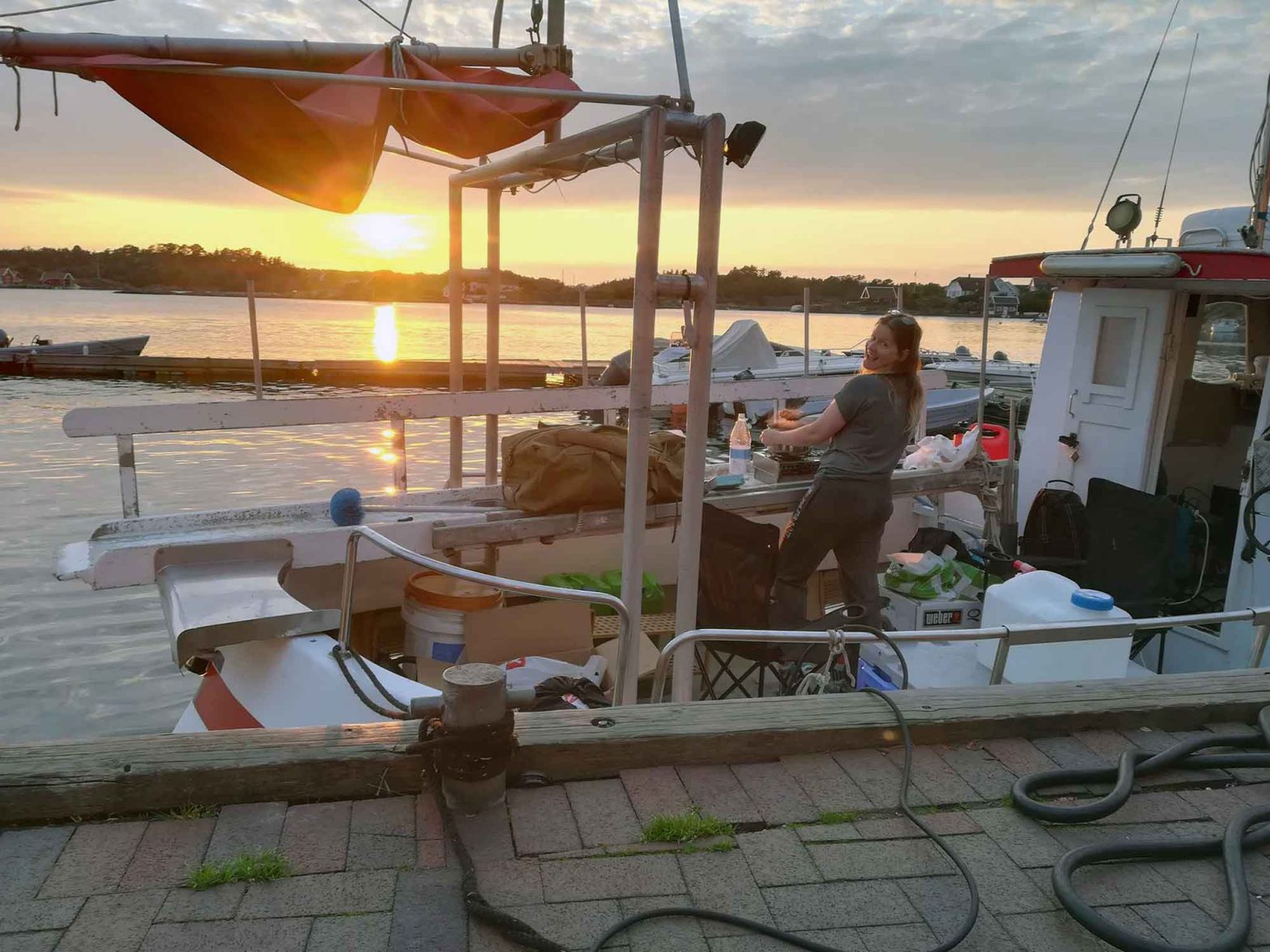 A boat with the sunset in the background and a woman cooking food on deck.