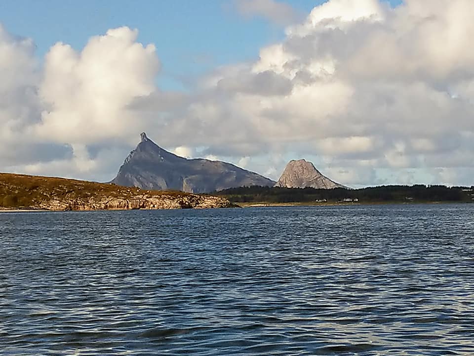 A pointy mountain in the distance with big clouds across the sky and the sea in the forefront.