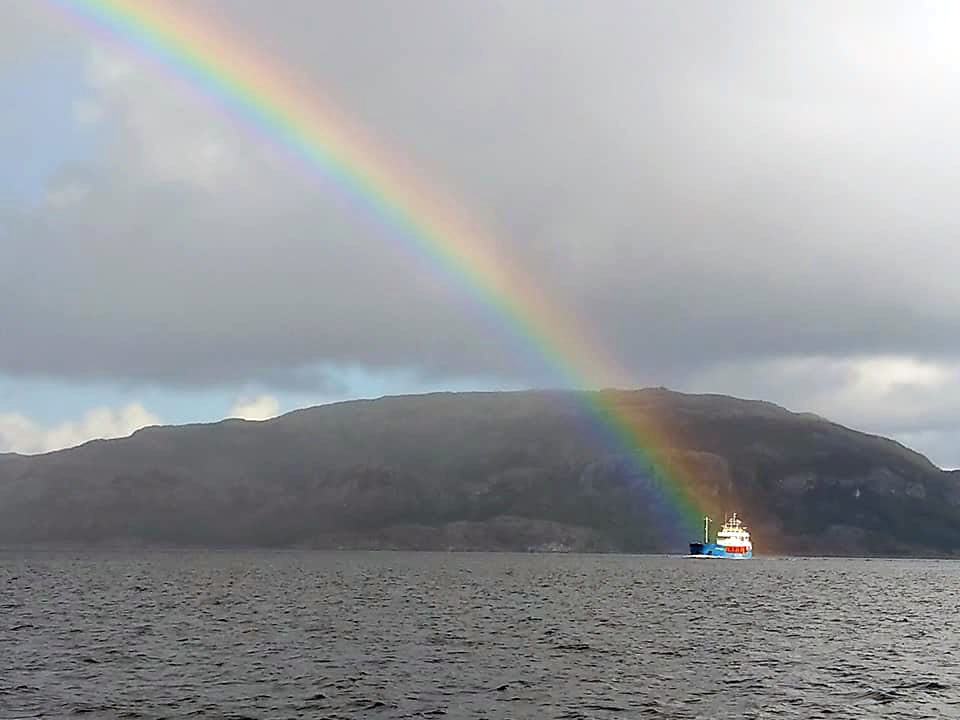 A rainbow that leads down onto a boat in the distance, with a big mountain in the background.