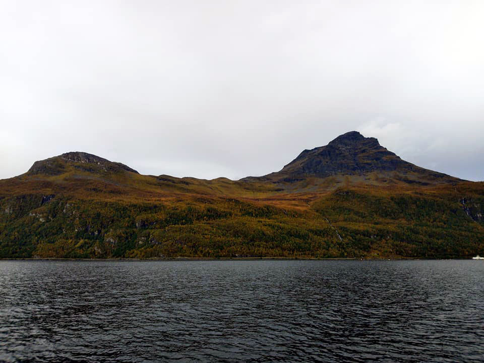 A mountain by the water covered in trees that are turning red and yellow in the fall weather.