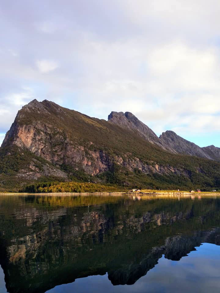 A wooded sloped mountain with extremely calm waters in front.