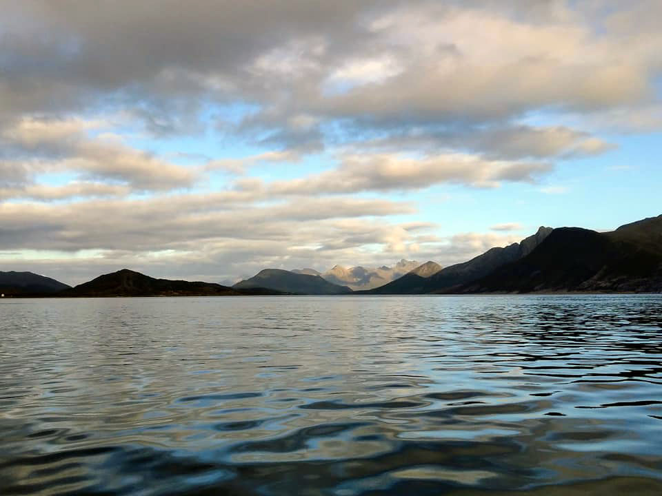 A long range of mountains with water in the forefront and a cloudy sky above.