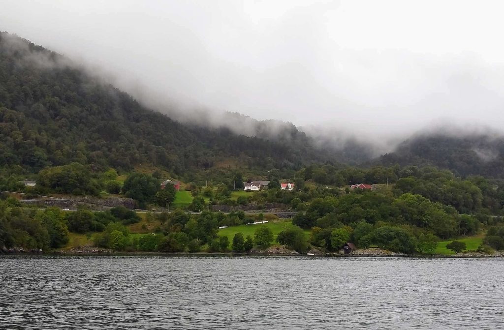 A house and a farm nested in the forest by the water, heavy clouds and mist overhead.