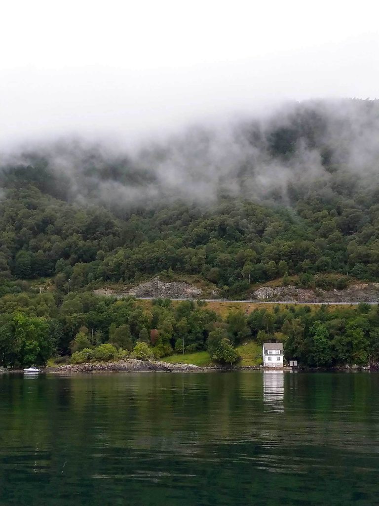 A wooded mountain with a single white house by the shore and mist rolling over the hill. 