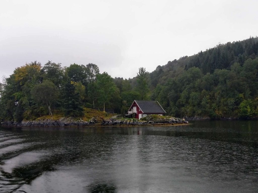 A small red cabin, right by the waters edge, surrounded by trees.