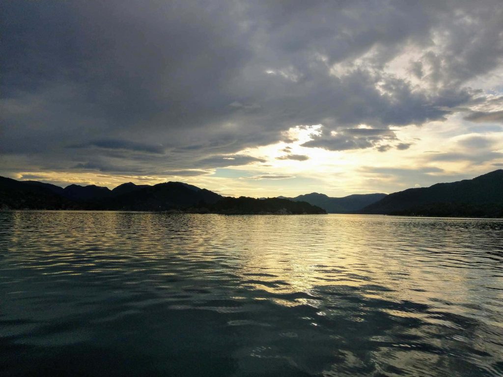 A dark image of water and a cloudy sky and the sun barely peaking through the clouds.