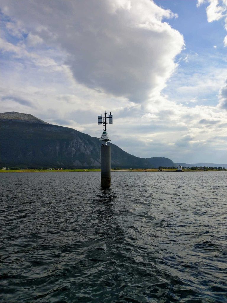 A guide post in the water with a mountain in the distance, and cloudy skies.