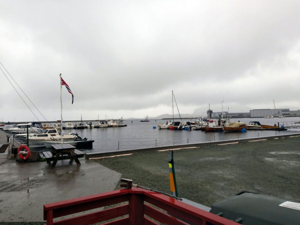 A marina with a park bench, a flag pole with the Norwegian flag and a few boats.