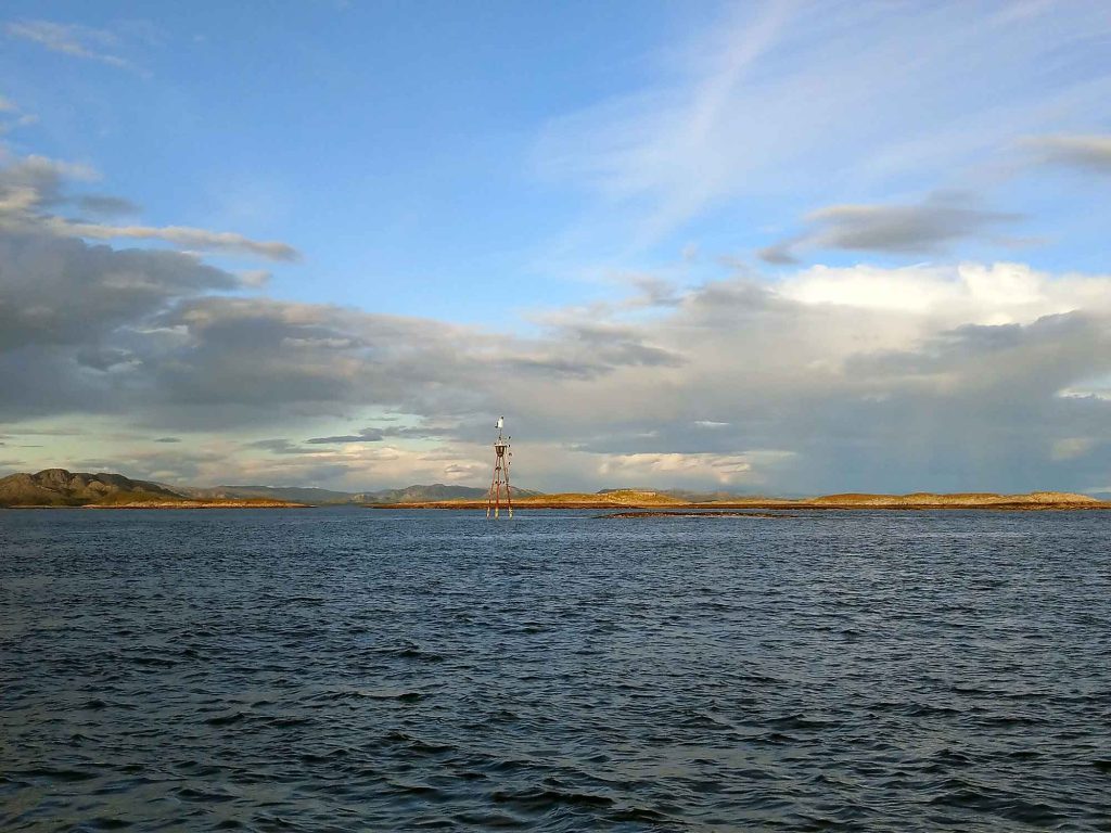 A shot of the water with a guide post in the middle, some flat islands and mountains in the distance.