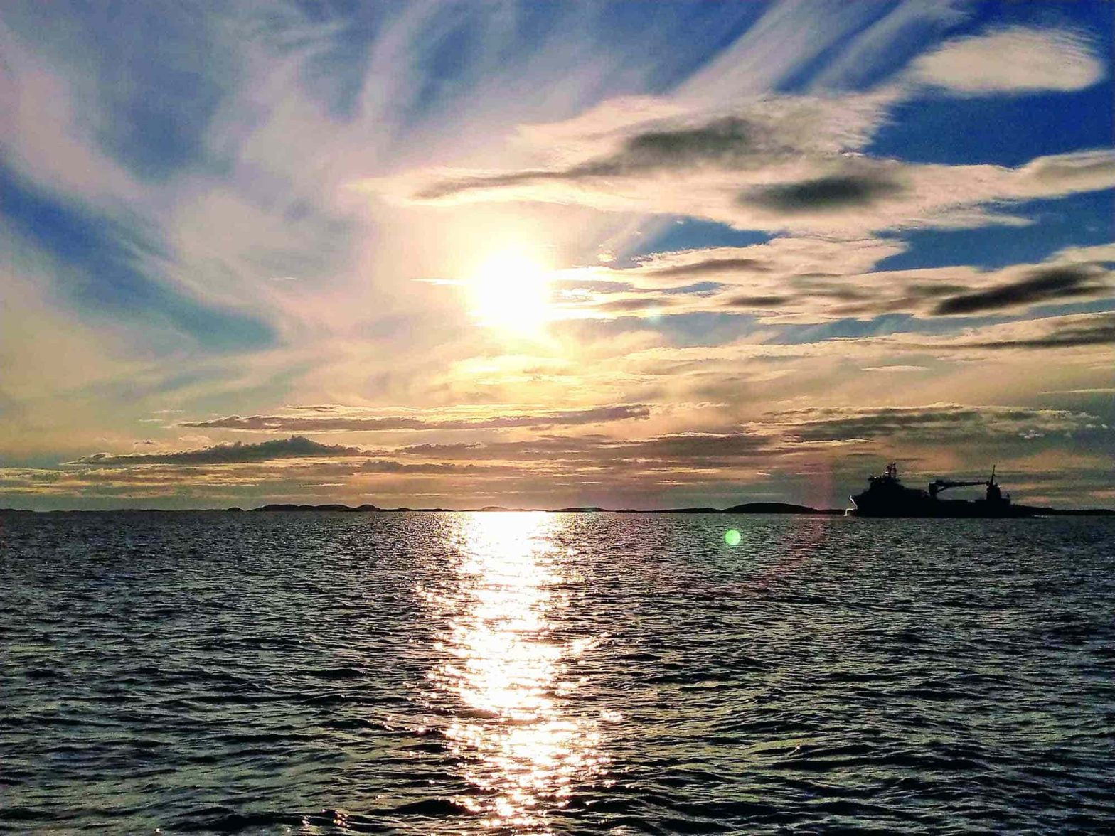 Calm sea with the sun shining brightly through whispy clouds and a ship passes in the horizon.