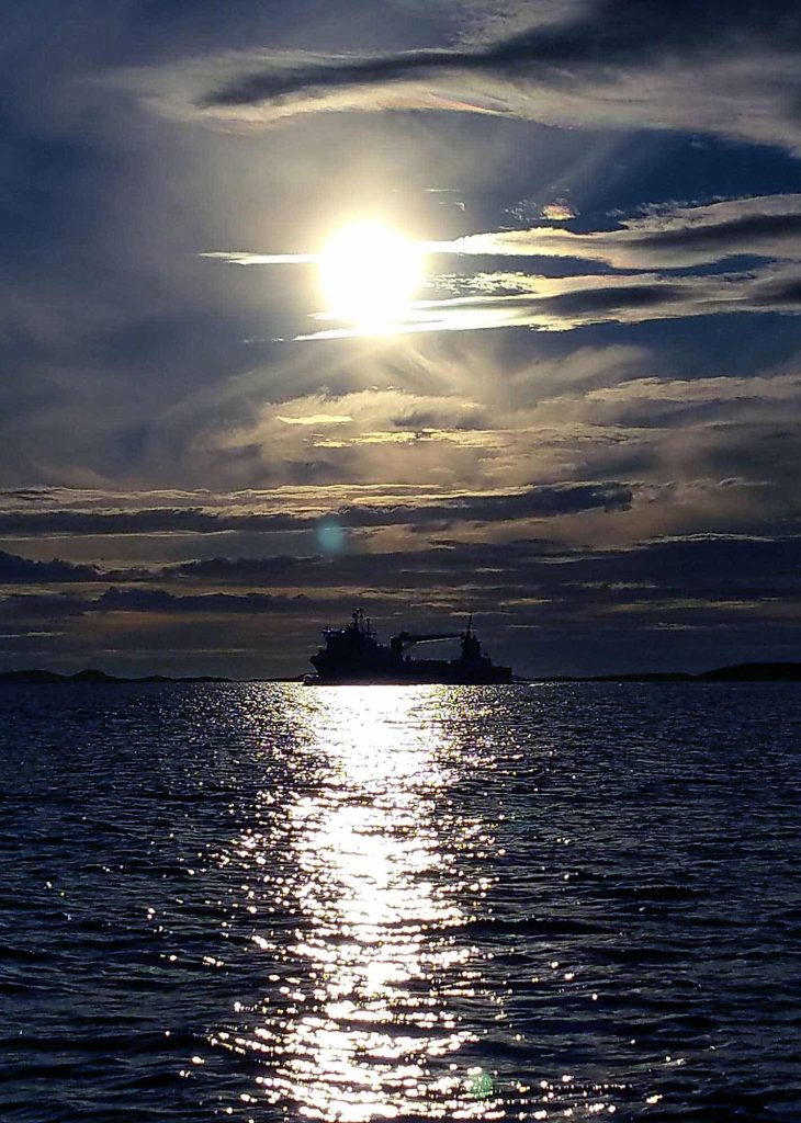 The silhouette of a ship in the distance, the sun shines brightly through the clouds and reflects in the water.